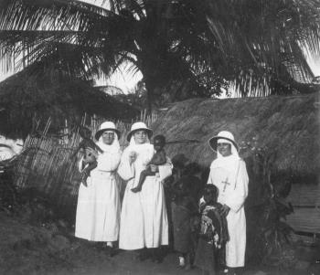 Drie missiezusters met een tropenhelm houden peuters op de arm vóór een huisje; Foto Foto