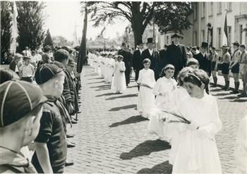 Op de dag van de viering van zijn 40-jarig priesterfeest loopt pastoor van Welie omrings door als bruidjes verklede meisjes van de pastorie naar de kerk. Langs de weg hebben zich verkenners en welpen opgesteld. Bijschrift: "De Blaathemseweg herschapen in een met zon overgoten landschap"; Foto Foto