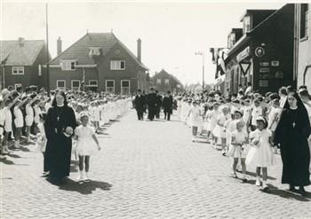 Op de dag van de viering van zijn 40-jarig priesterfeest loopt pastoor van Welie omringd door bruidjes naar de kerk. Langs de weg staan kinderen in gymnastiekpakken die de pastoor groeten. Bijschrift: "IJverige zusters begeleiden de talloos vele bruidjes"; Foto Foto