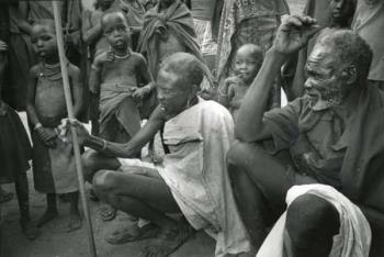 Uganda/Twee hurkende oudere mannen daar omheen kinderen en volwassenen.; Foto Foto