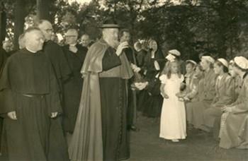 Het apostolaat der Hereniging heeft een kardinaal Tisserant uitgenodigd. De kardinaal zegent hier een groep jonge vrouwen in het park van de assumptionisten. De priester achter de bisschop, die zijn hand bij zijn boord houdt, is de voorzitter van het Apostolaat der Hereniging, pastoor A.G. Smit. De foto's 6a1455, 6a1456, 6a1457 en 6a1458 hebben allemaal betrekking op het bezoek van de kardinaal aan Nederland; Foto Foto