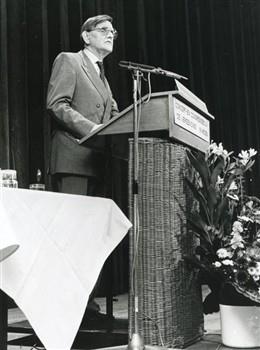 Kees Fens staat achter de microfoon ter gelegenheid van het 65-jarig bestaan van de universiteit, met tekst 'concert- en congresgebouw De Vereeniging Nijmegen'; Foto Foto
