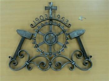 Metalen klok in de vorm van een rad, twee hamers en een kruis, met tekst: '1903-1953'; Klok Klok