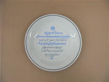 Bord houdende het embleem van de RK Vereniging Handenarbeid, met tekst: 'Het 300ste Examen Creatieve Handvaardigheid A-gedeelte werd op 8 maart 1969 door de R.K. Ver. 'Handenarbeid' afgenomen te Gemert in de R.K. Levensschool Gerardusplein 2'.
Achterkant houdende het stempel van Sphinx, Maastricht, met tekst: 'Royal Sphinx Maastricht P. Regout Made in Holland porselein'; Gedenkbord Gedenkbord