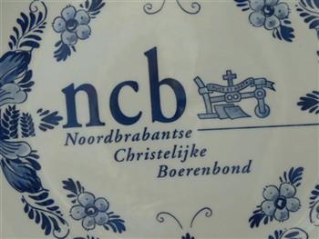 Wandbord, met embleem van de Noord-Brabantse Christelijke Boerenbond, houdende afbeeldingen van bloemen, een koe, een varken en een kip. Op achterzijde tekst 'Koos bedankt, voor 31 jaar bestuurlijk werk NCB Velddriel' en 'Delft Plateelbakkerij Schoonhoven Holland B.'; Wandbord Wandbord