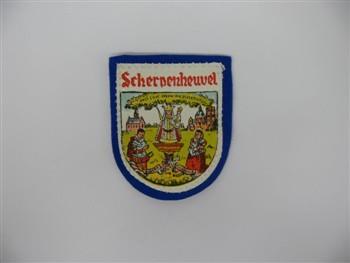 Schildvormige badge houdende een afbeelding van Onze Lieve Vrouwe van Scherpenheuvel met links van haar een kerk met daaronder een knielende edelman 'Albert' en rechts van haar een basiliek (basilica minor) met daaronder een knielende edelvrouw 'Isabella'. Met tekst: 'Scherpenheuvel'; Badge Badge